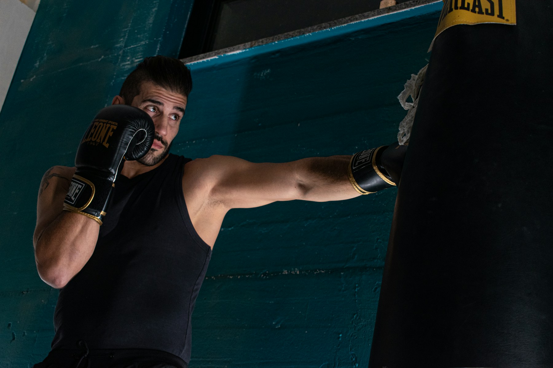 Full-color photo of one man in black and gold boxing gloves throwing a punch at a heavy bag against a teal wall