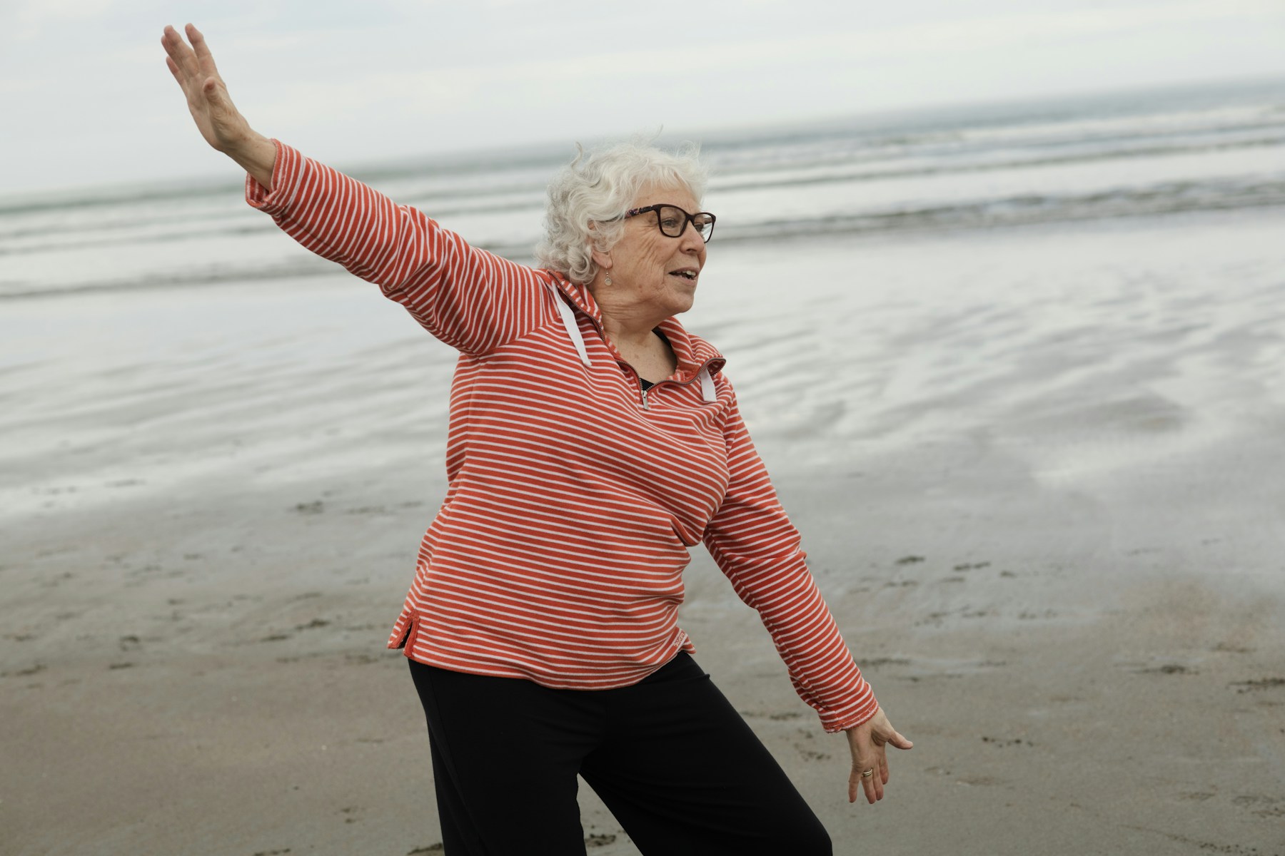 Full-color photo of a visibly older woman with gray hair and glasses in a striped top, alone on a beach with one arm stretched overhead