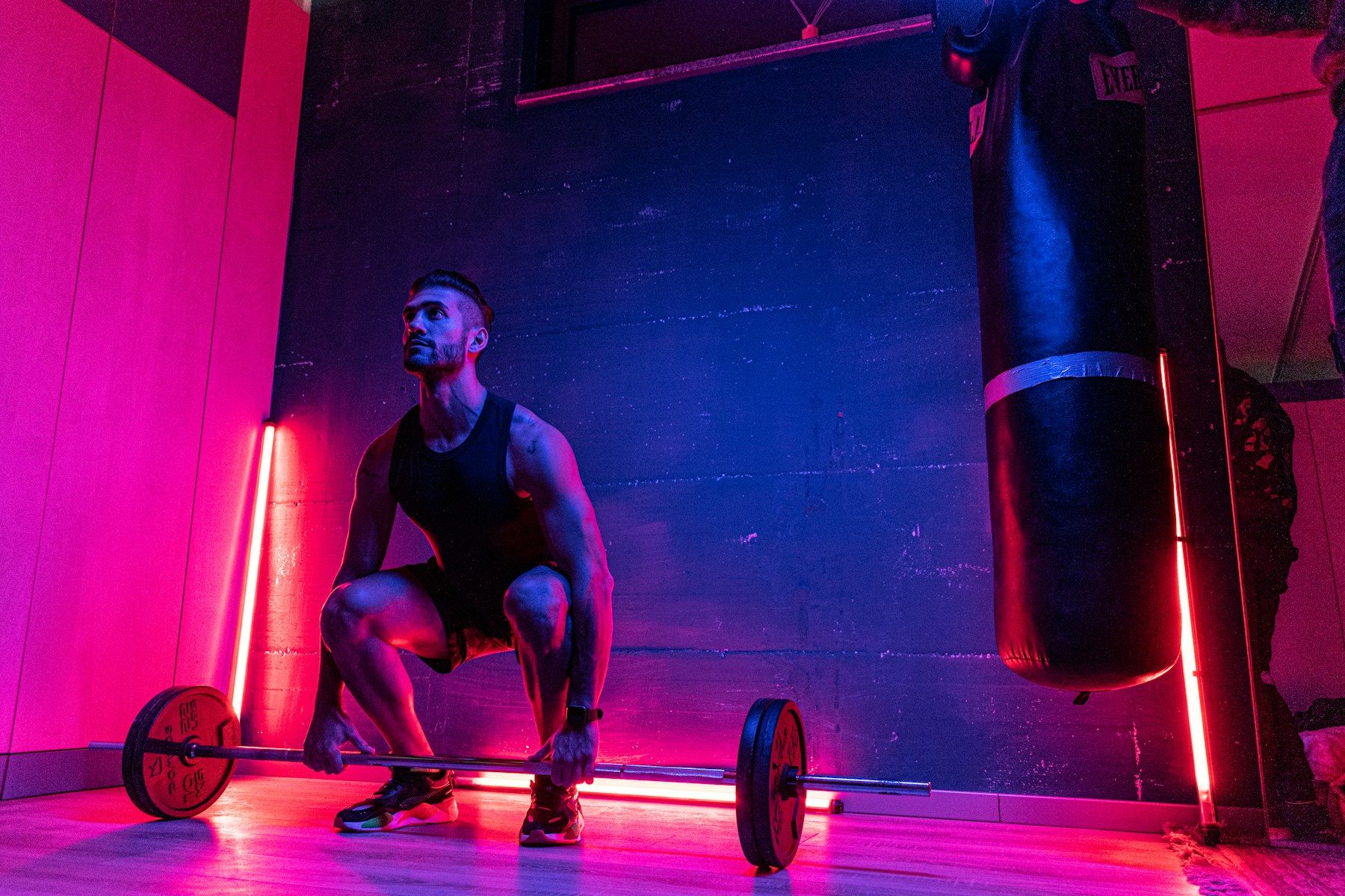 Full-color photo of one man in a crouched deadlift setup gripping a loaded barbell in a gym lit with pink and blue light